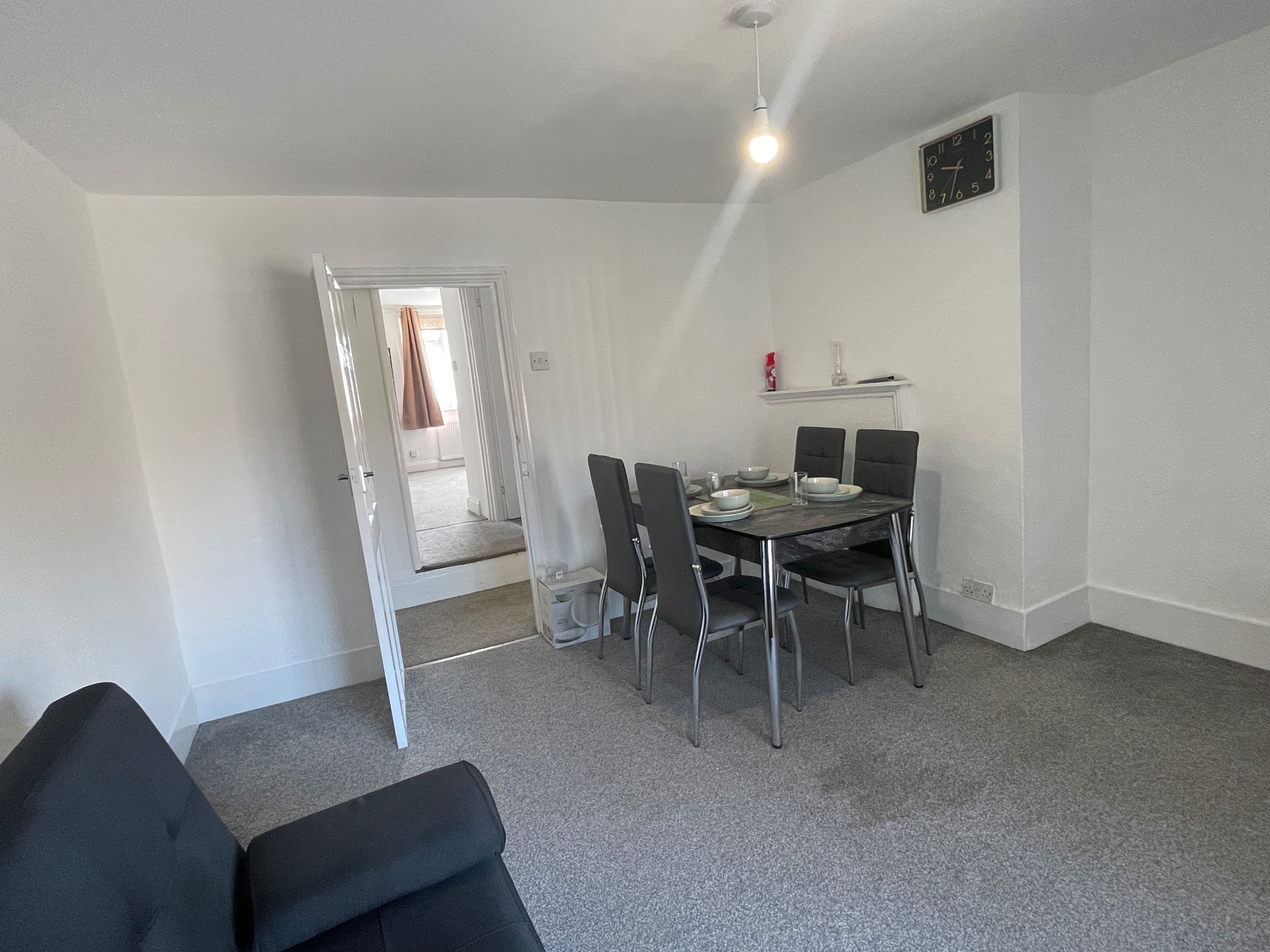 Een eetruimte met een tafel voor vier personen, vier stoelen, een klok aan de muur en een deur naar een andere kamer.