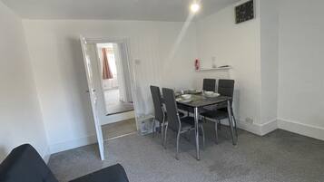 Een eetruimte met een tafel voor vier personen, vier stoelen, een klok aan de muur en een deur naar een andere kamer.