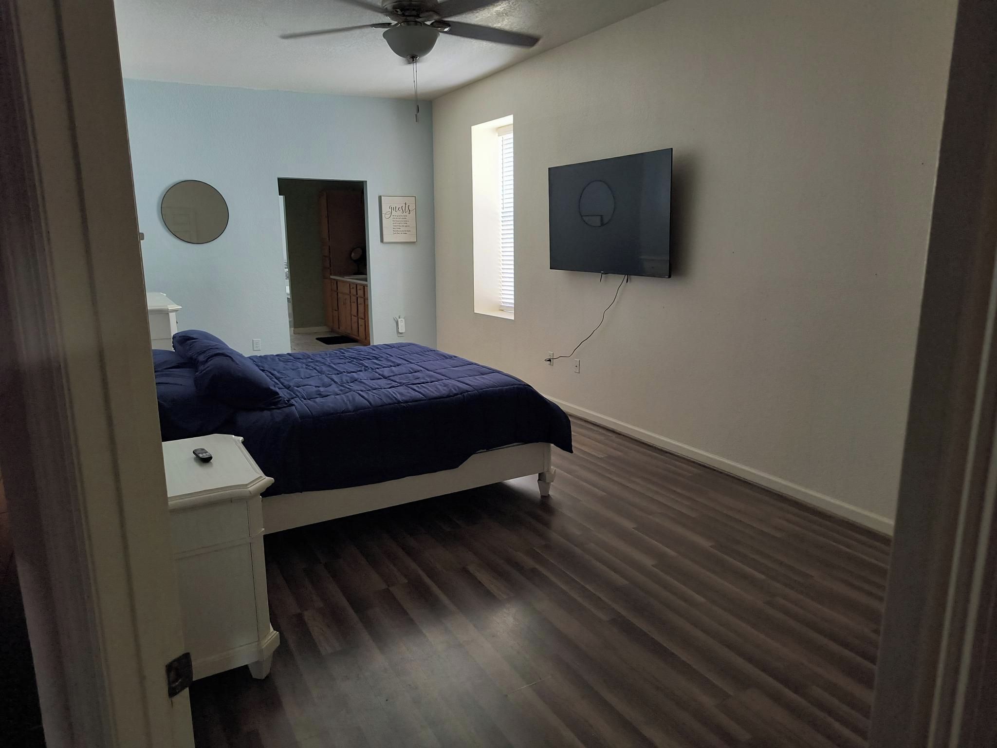 A bedroom with a bed, a nightstand, a ceiling fan, a window with blinds, and a wall-mounted TV.