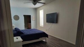 Un dormitorio con cama, mesita de noche, ventilador de techo, ventana con persianas y televisor montado en la pared.