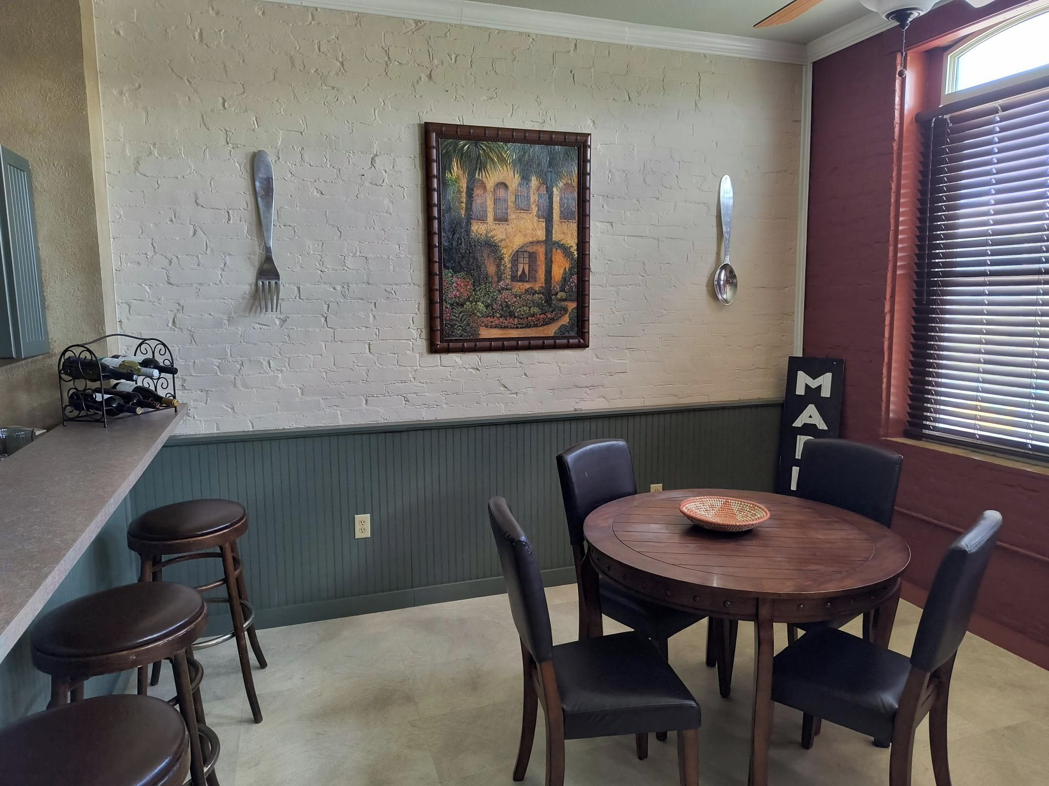 A dining area with a round table, four chairs, a painting on the wall, and a window with blinds.