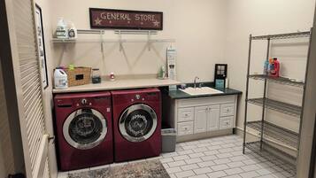 A laundry room with two front-loading washers, a sink, and shelves with cleaning supplies.
