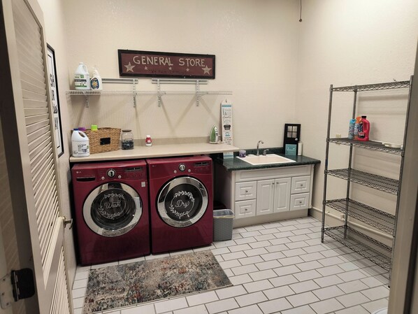 A laundry room with two front-loading washers, a sink, and shelves with cleaning supplies.