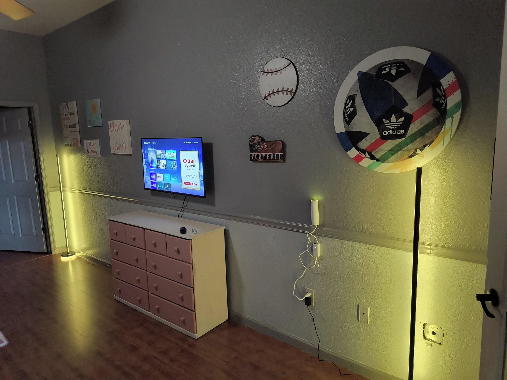 A room with a television mounted on the wall displaying a streaming service interface, a shoe-shaped football sticker, and a circular Adidas logo.
