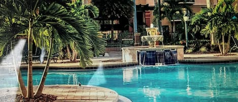 Una piscina con una fuente, rodeada de palmeras y un edificio con balcones.