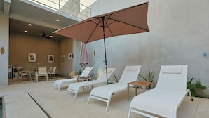 Uma área de estar interna e bem iluminada pelo sol, com poltronas brancas, uma mesa, cadeiras e um ventilador de teto.