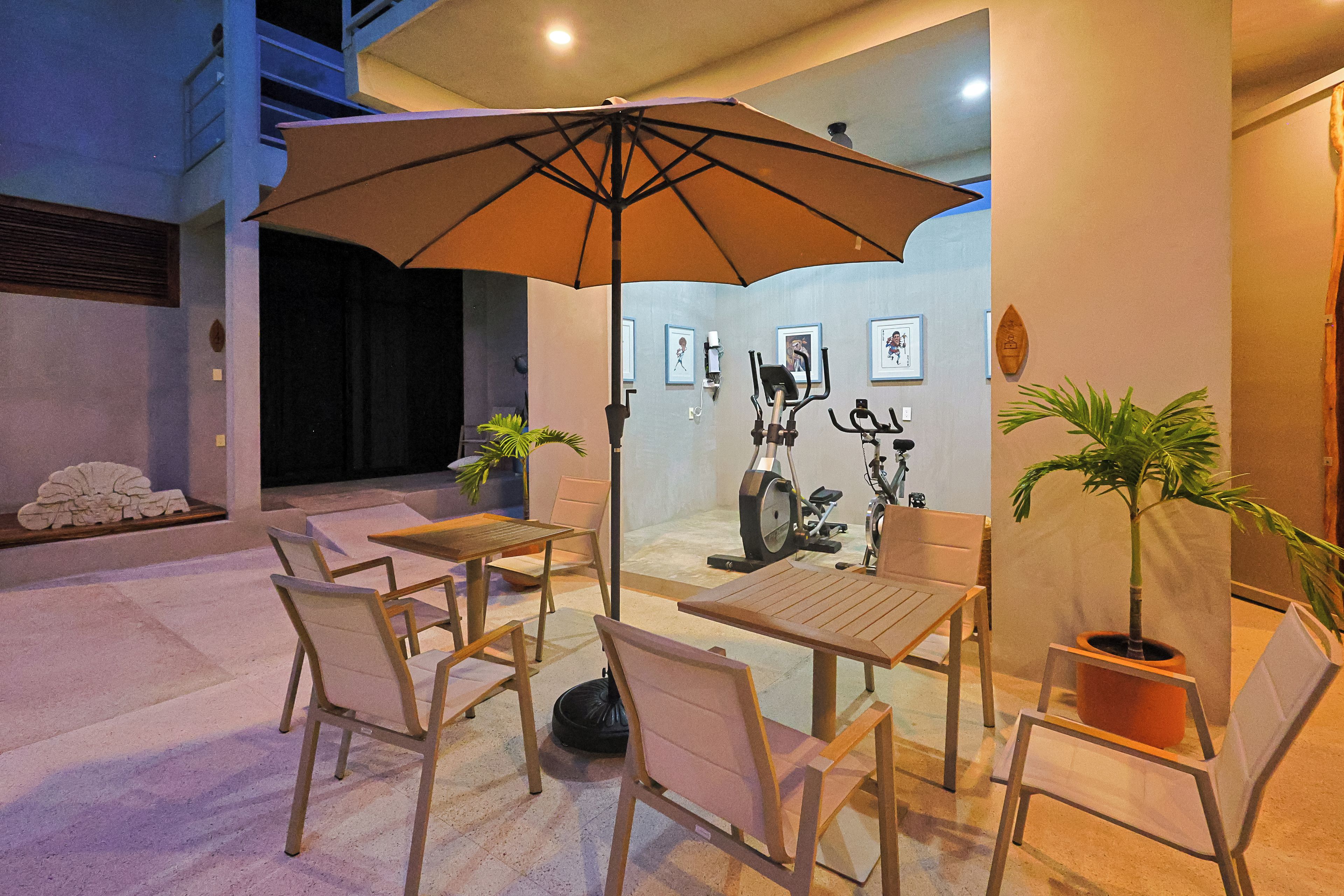 An indoor seating area with tables and chairs, an umbrella, exercise equipment, and artwork on the walls.