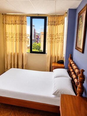A hotel room with a bed, curtains, a window, a nightstand, and a wall-mounted picture. - HOTEL MILENIO (Guayaquil)