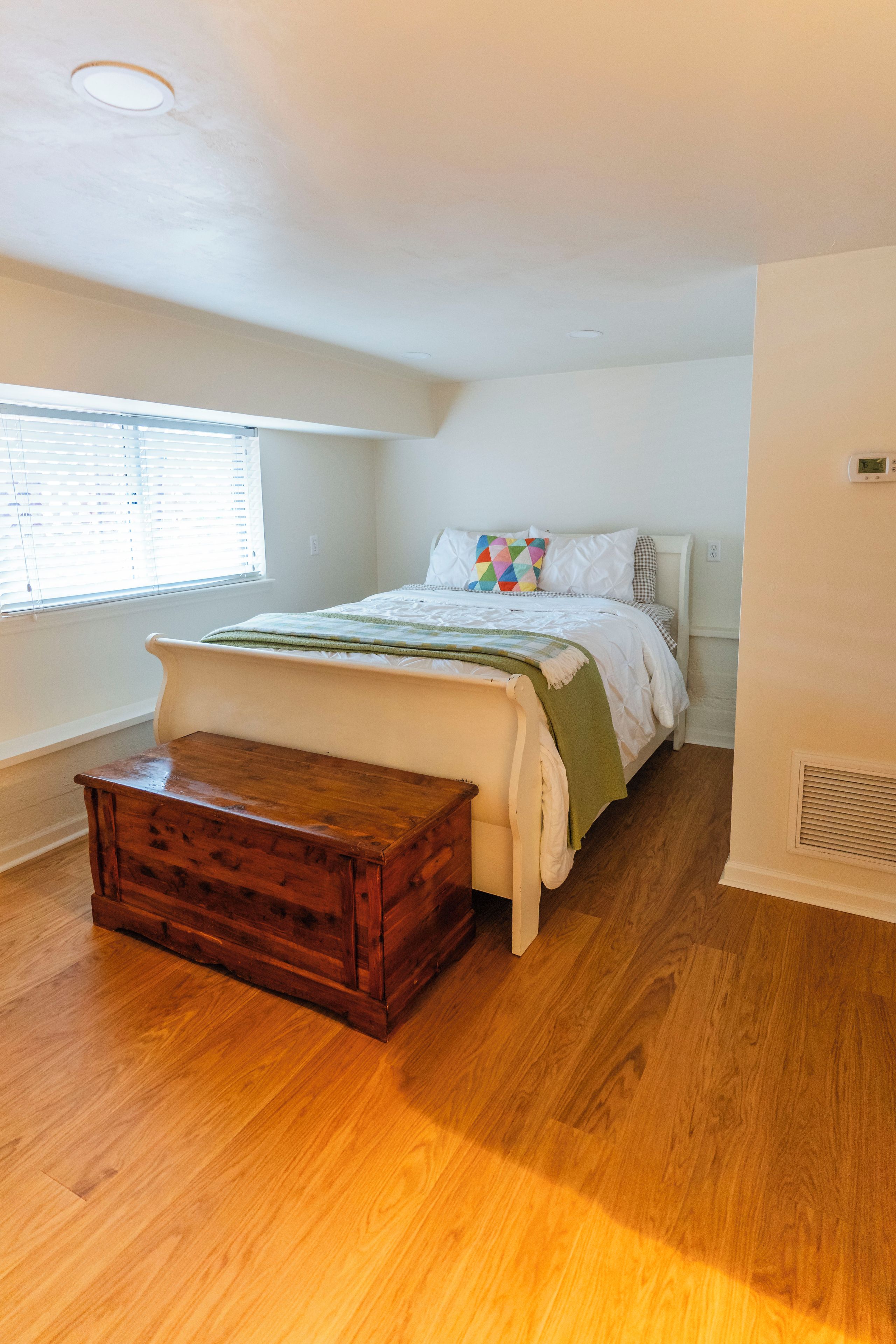 A bedroom with a wooden bench, a bed with a colorful pillow, and a window with blinds.