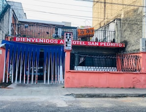 A hotel entrance with a red and blue gate, a sign reading 'BIENVENIDOS A HOTEL SAN PEDRITO,' and a 'HOTEL' sign above the gate.