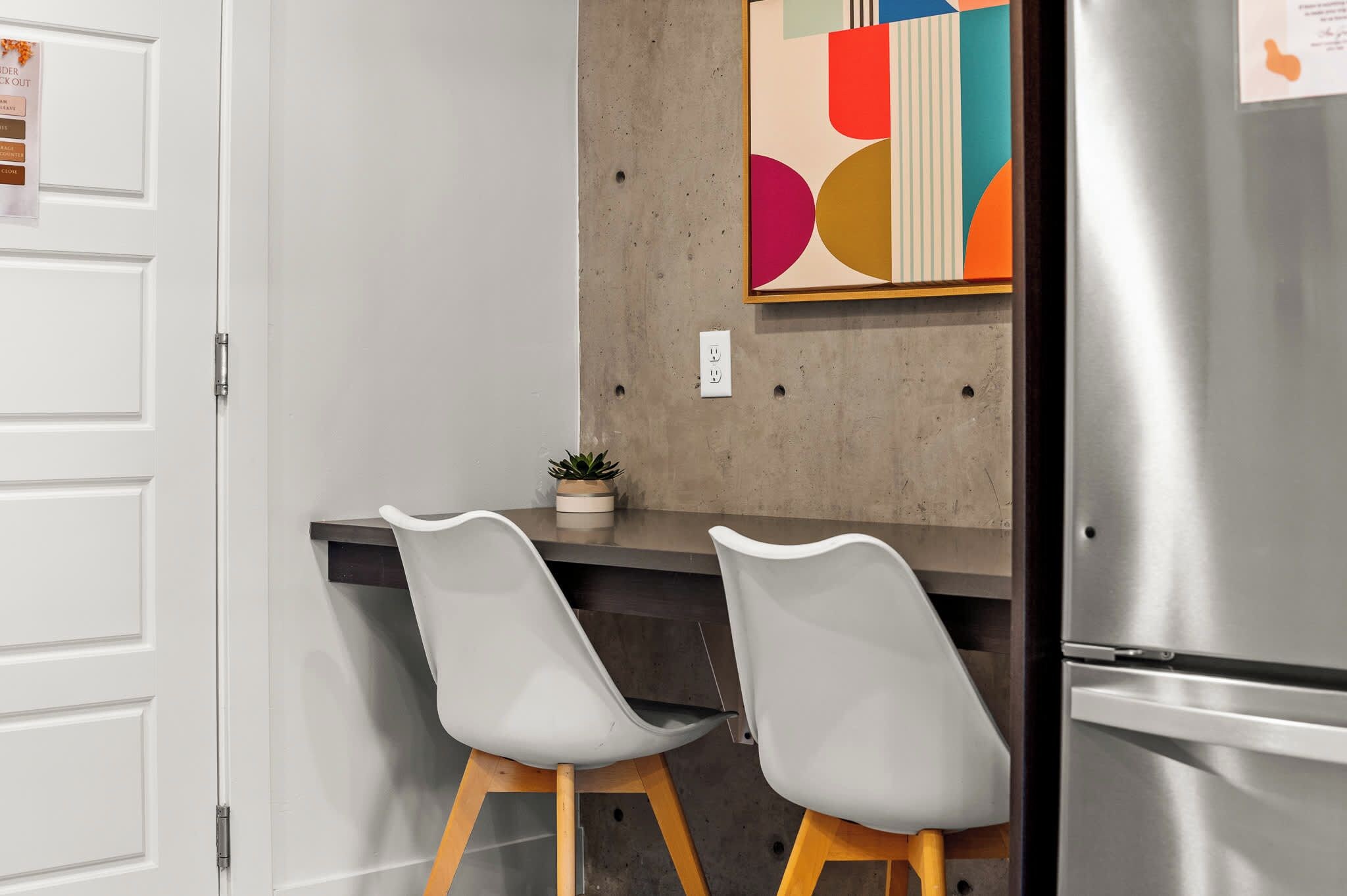 A modern kitchen with two white bar stools, a stainless steel refrigerator, and a colorful abstract wall art piece.