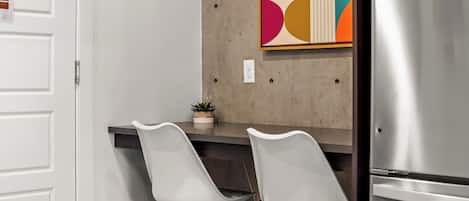 A modern kitchen with two white bar stools, a stainless steel refrigerator, and a colorful abstract wall art piece.