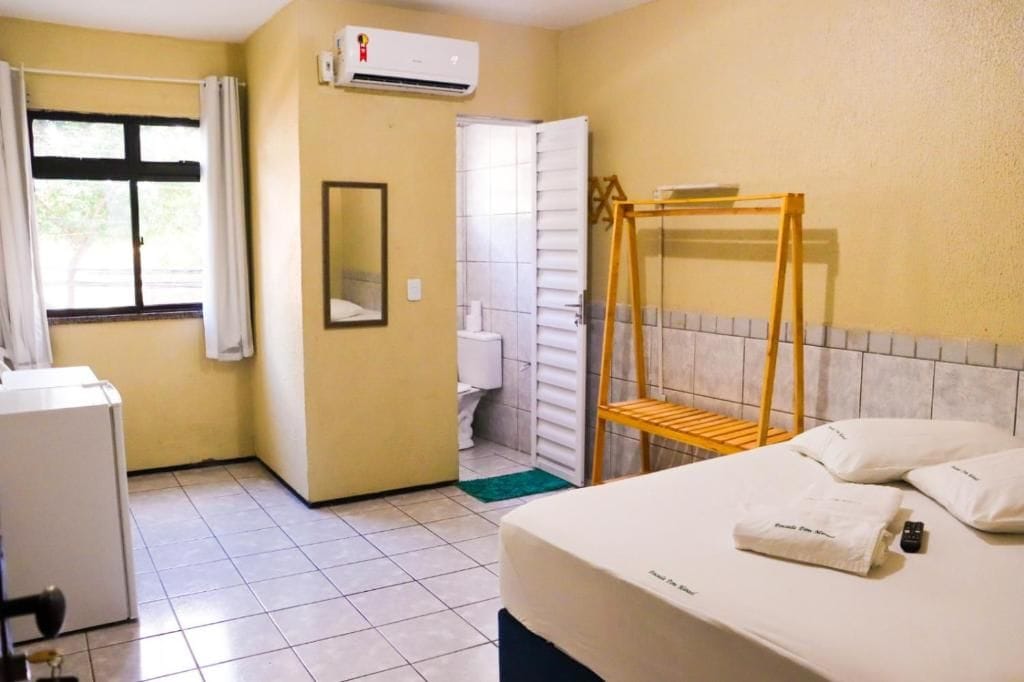 A hotel room with a bed, a wooden bunk bed, a window with curtains, an air conditioning unit, a mirror, and a small table with a remote control.