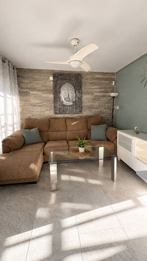 Un salon moderne avec un canapé d’angle, une table basse en verre, une statue de Bouddha, un ventilateur de plafond et une horloge murale.