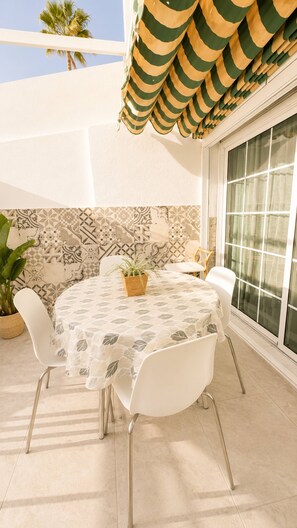 An outdoor patio with a round table, white chairs, a potted plant, and a patterned wall. - Sonnenland Bungalow · 2 bedrooms · 4 people · Terrace & Pool (Maspalomas)