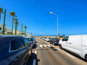 Een kustweg met geparkeerde auto's, een witte bus en een strakblauwe lucht.