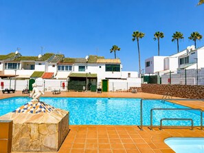 A swimming pool with a tiled deck, a statue, and a row of white buildings with green roofs and palm trees. - Sonnenland Bungalow · 2 bedrooms · 4 people · Terrace & Pool (Maspalomas)