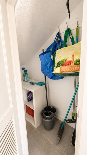 A corner of a room with a white shelf, a grey dustpan, a broom, and a blue bag hanging on a hook. - Sonnenland Bungalow · 2 bedrooms · 4 people · Terrace & Pool (Maspalomas)