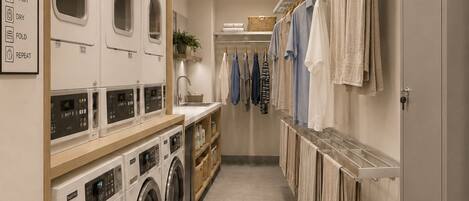 A laundry room with multiple washing machines, a drying rack, and a shelf with folded clothes.