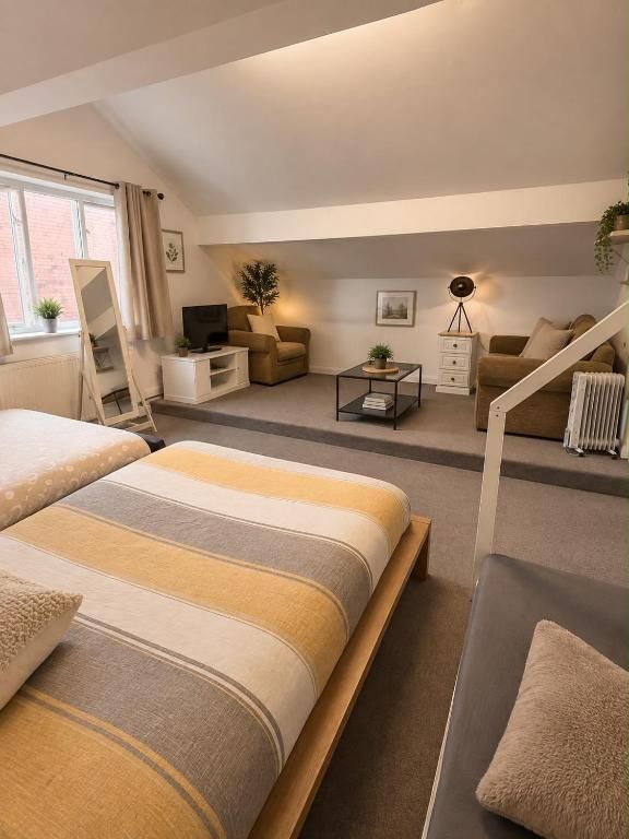 Una habitación de hotel moderna con una cama grande, un sofá, un escritorio y una silla. Cuenta con una ventana con cortinas, un radiador y una planta en el alféizar.