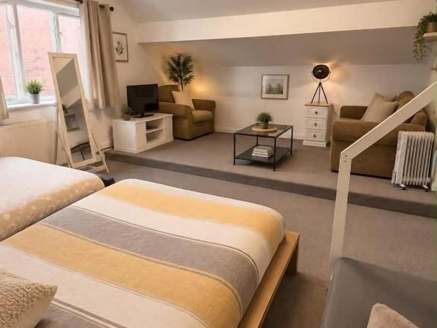 Una habitación de hotel moderna con una cama grande, un sofá, un escritorio y una silla. Cuenta con una ventana con cortinas, un radiador y una planta en el alféizar.