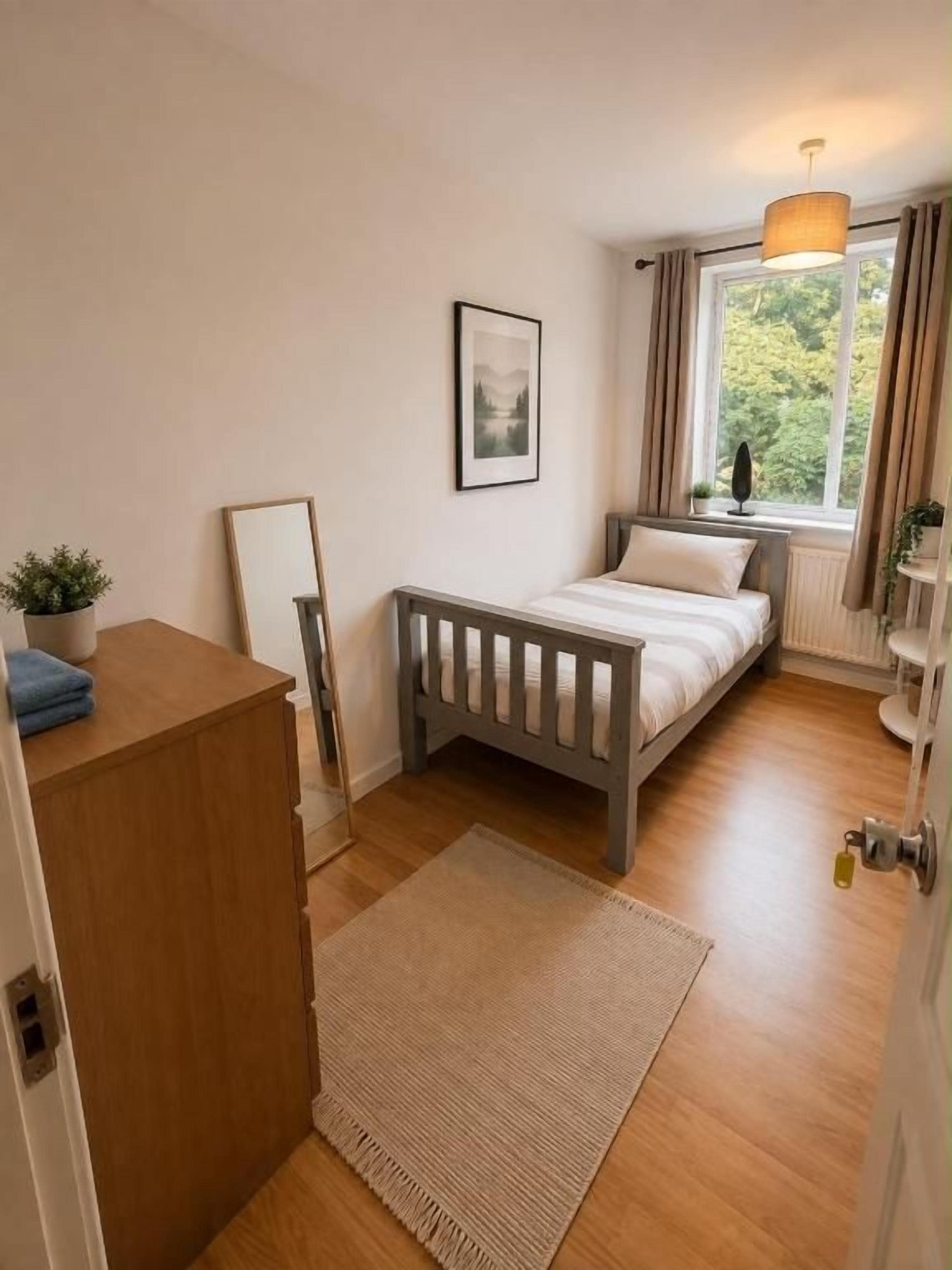 Une chambre à coucher avec un lit, une table de chevet, une lampe, une fenêtre et une vue sur la verdure à l’extérieur.