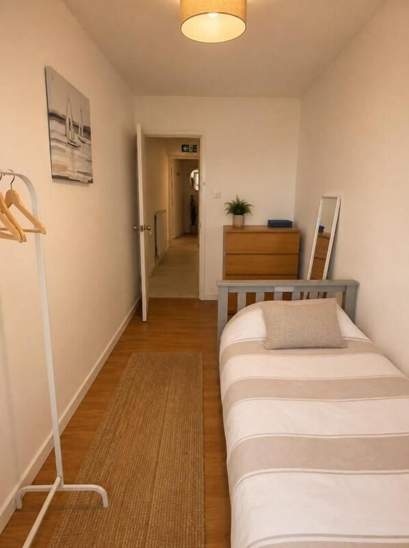 Une chambre d’hôtel étroite, comprenant un lit, une commode, un miroir, un porte-manteau mural et une porte donnant sur un couloir.