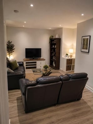 Una sala de estar moderna con un sofá de cuero oscuro, un televisor de pantalla plana, una mesa de centro de madera y una estantería.