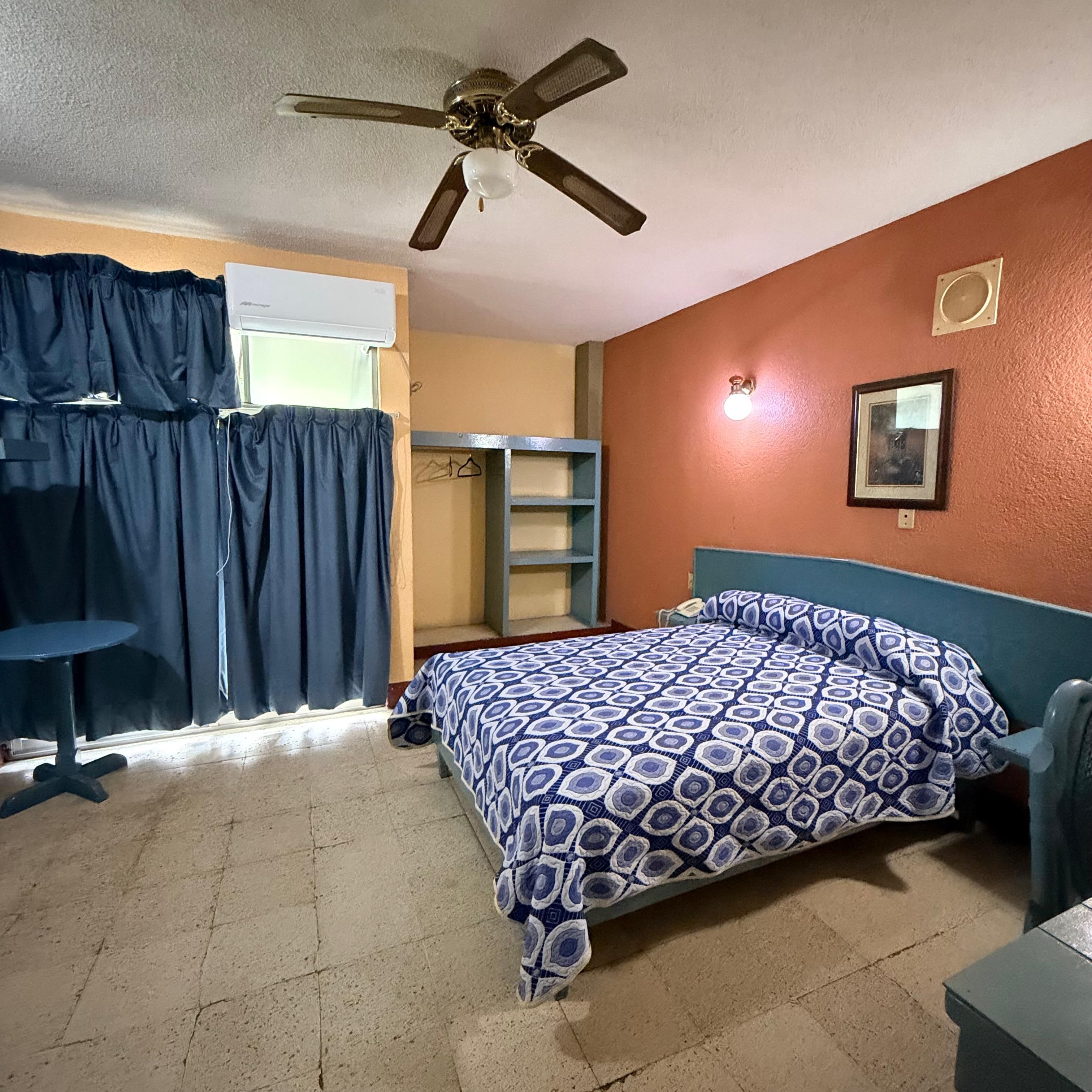 Un dormitorio con cama, escritorio, silla, ventilador de techo, ventana con cortinas y estante en la pared.