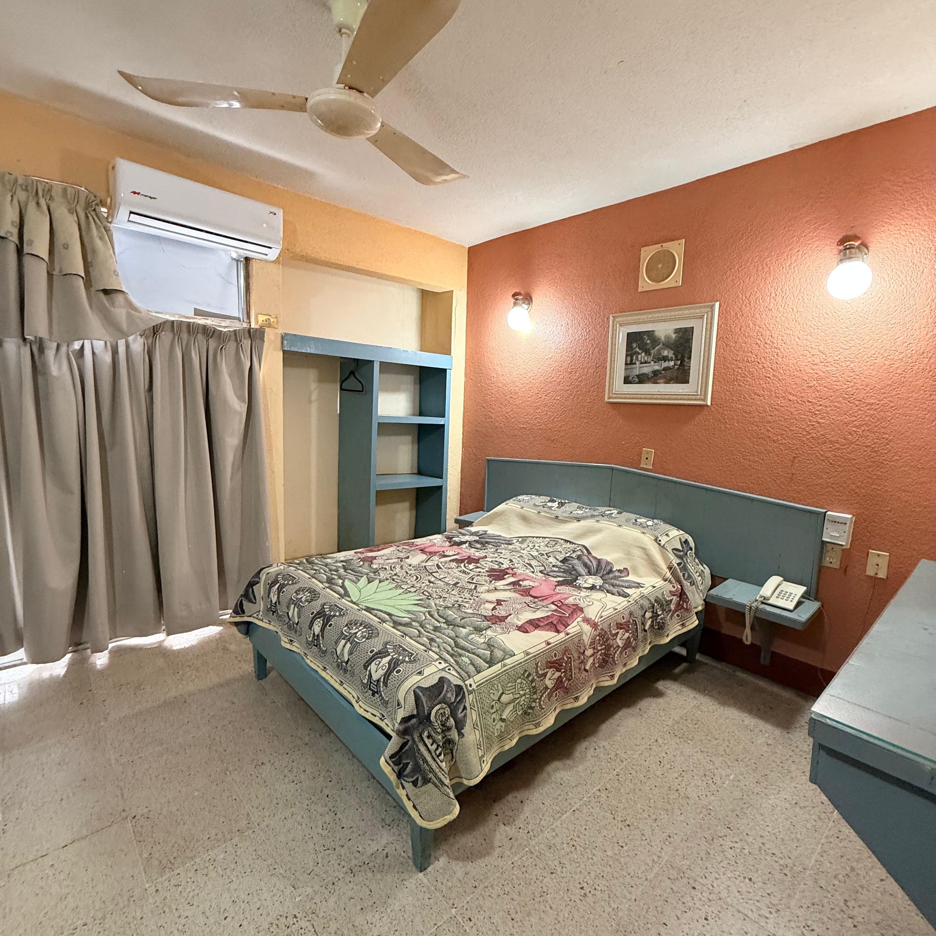 A bedroom with a bed, an air conditioning unit, a ceiling fan, a bedside telephone, and a wall-mounted picture.