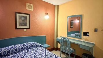 A hotel room with a bed, a desk, a chair, a mirror, and a trash bin.