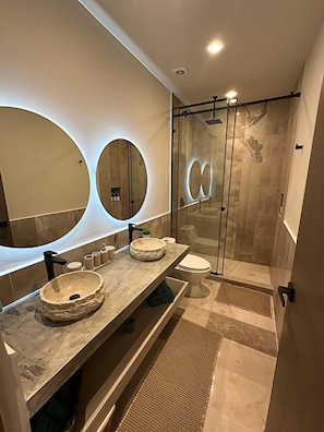 Un baño moderno con un espejo grande, dos lavamanos con cuencos redondos, un inodoro y una cabina de ducha con paredes de vidrio.