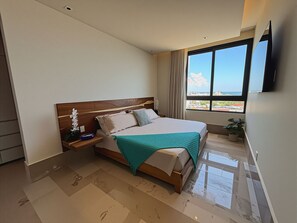 Habitación de hotel moderna con una cama grande, una mesita de noche, una planta en maceta y vista a la ciudad a través de un amplio ventanal.