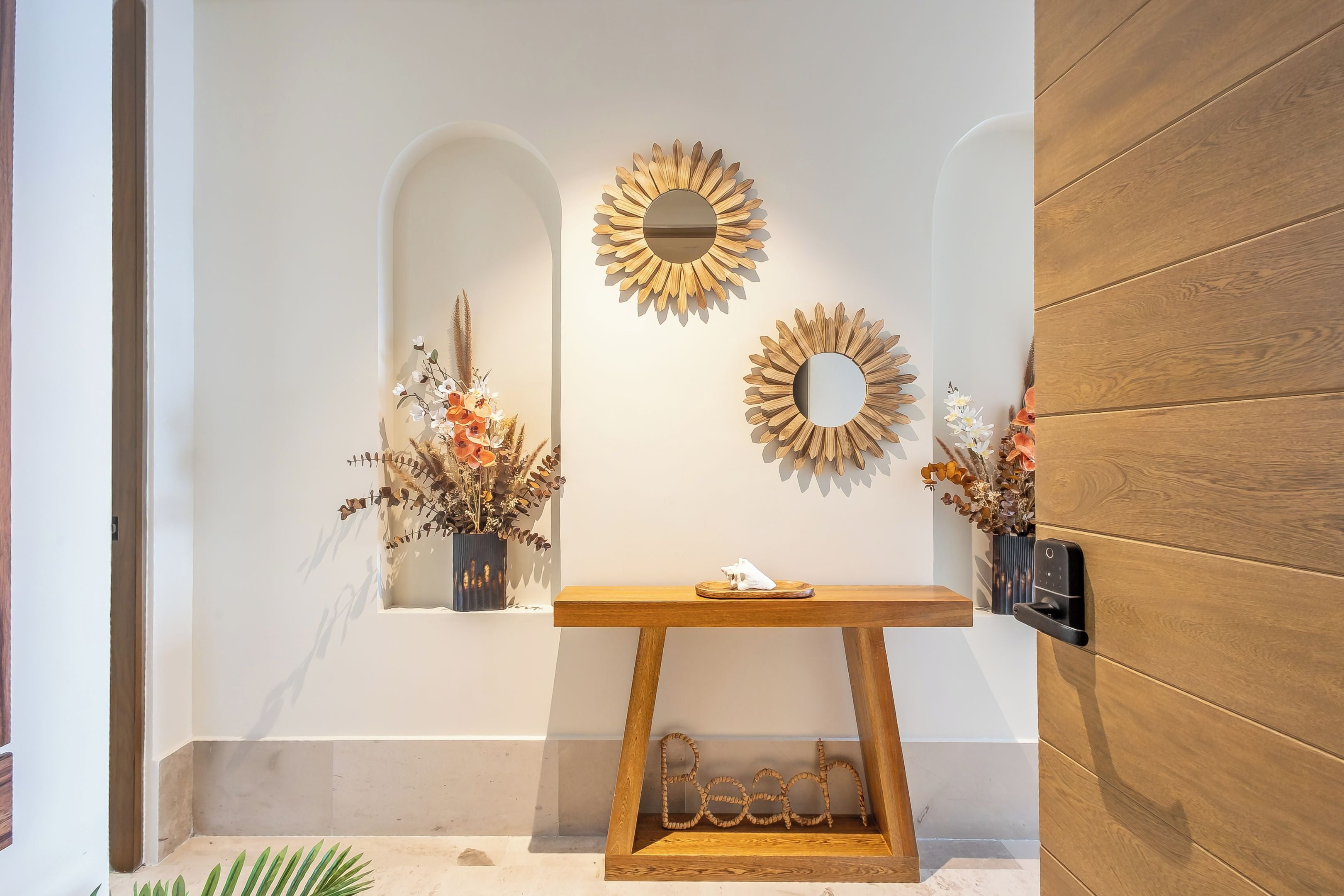 Uma área de recepção com um aparador de madeira, dois espelhos fixados na parede e arranjos florais decorativos.