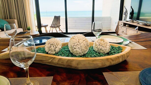 Ein modernes Wohnzimmer mit einem Holzesstisch, zwei Weingläsern, einem dekorativen Mittelstück und Blick aufs Meer durch große Fenster.