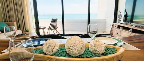 A modern living room with a wooden dining table, two wine glasses, a decorative centerpiece, and a view of the ocean through large windows.