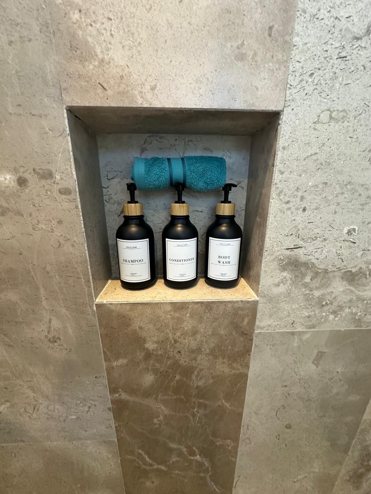 Un estante en el baño con tres botellas etiquetadas como 'Shampoo', 'Acondicionador' y 'Gel de ducha'.