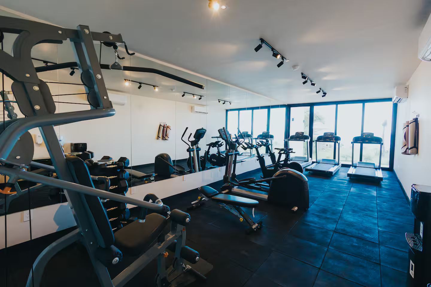 Uma academia com diversos equipamentos de exercício, amplas janelas e piso azul.