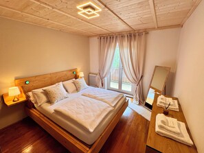 Un letto matrimoniale con testiera in legno, lampade ai comodini, una finestra con tende, un comò in legno con asciugamani e uno specchio.