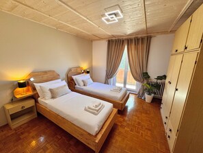 A bedroom with two wooden beds, a nightstand with a lamp, a potted plant, and a view of the outdoors through a window with curtains. - Terrific 2-bedroom apartment with AC in Trentino-Alto Adige (Trentino-Alto Adige)