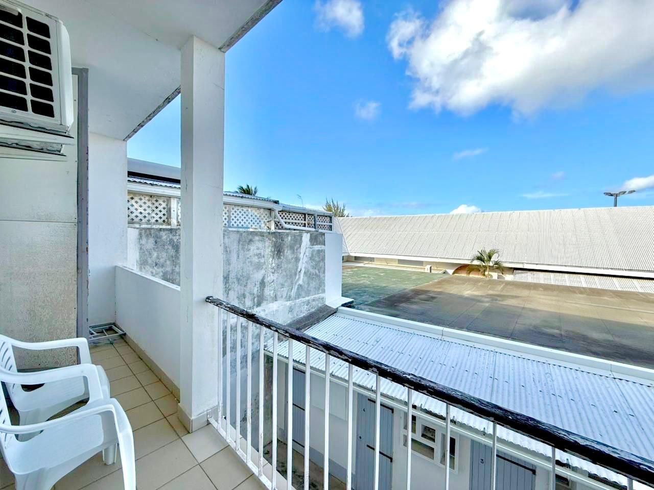 A balcony with a metal railing, white plastic chairs, and a view of a parking area and buildings in the distance.