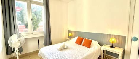 A hotel room with a bed, two bedside lamps, a fan, and a window with curtains.