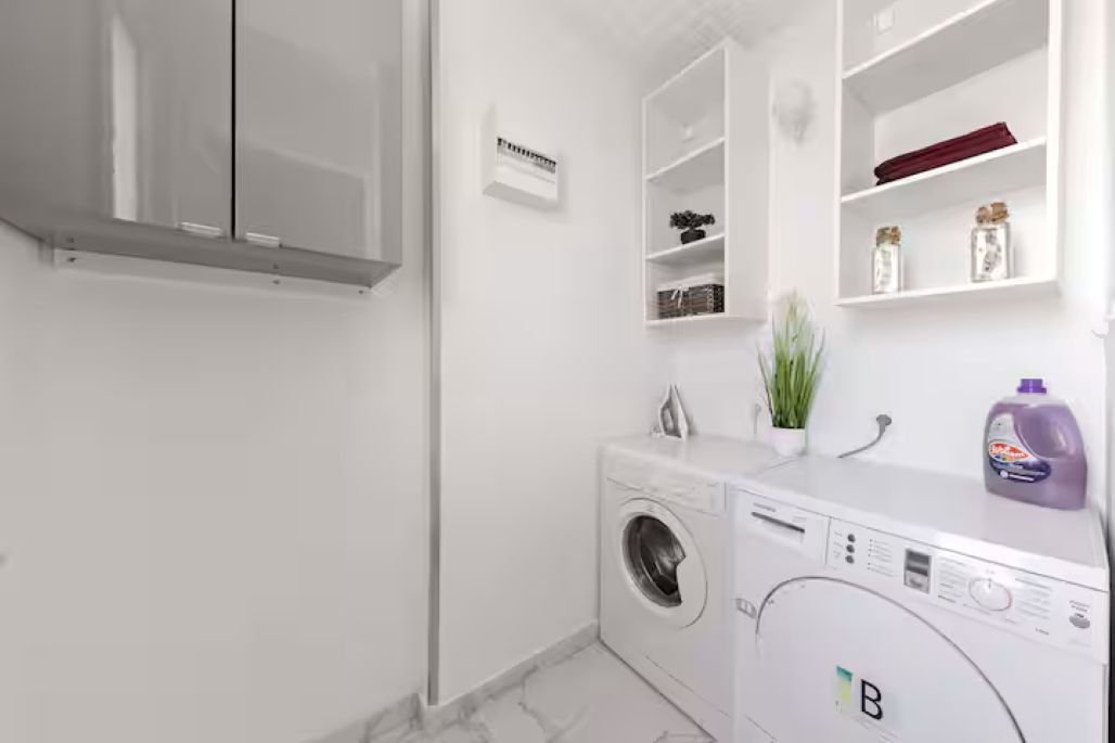 Une petite buanderie équipée d’un lave-linge à chargement frontal, d’un étendoir et d’une étagère contenant des produits de nettoyage.