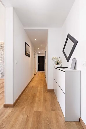 Un pasillo con suelo de madera, paredes blancas, un cuadro enmarcado y un pequeño armario blanco con un jarrón de flores.