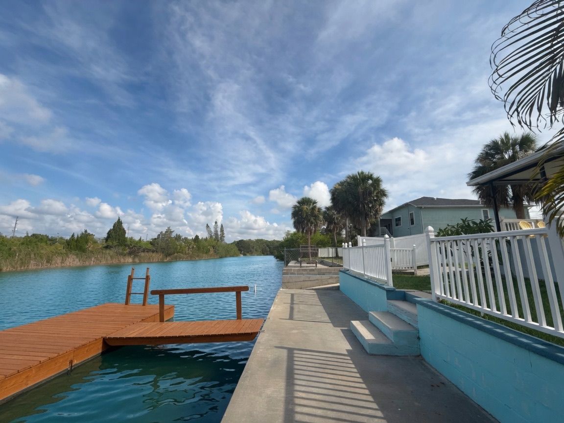 A wooden dock extending into a calm body of water, a concrete pathway alongside, and a white fence with steps leading to a house.