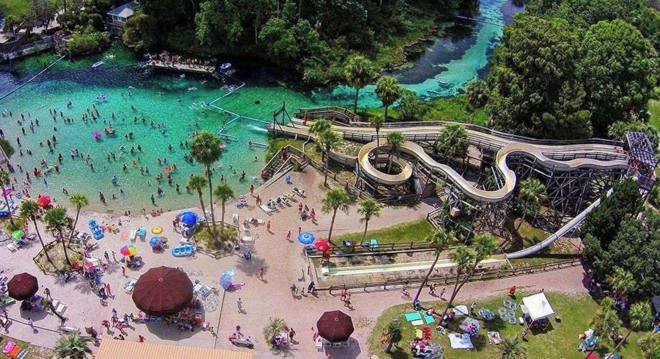 An aerial view of a beach with a water slide, clear turquoise water, and people enjoying the area.