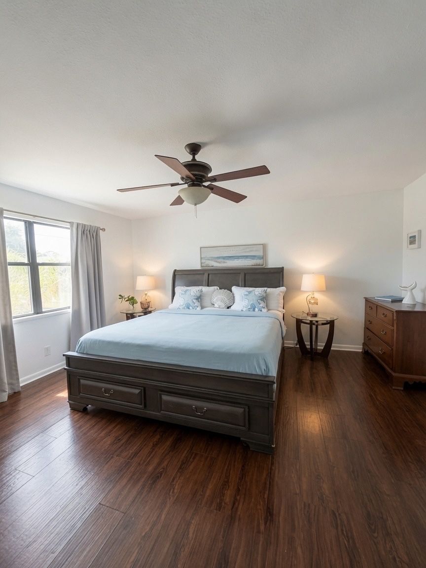 Une chambre à coucher avec un lit, des lampes de chevet, un ventilateur de plafond, une commode et une fenêtre avec des rideaux.