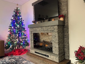 A living room with a fireplace, a Christmas tree, and a flat-screen TV mounted above the fireplace.