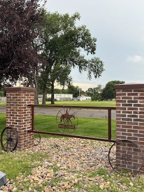 A metal gate with a horse and wagon wheel emblem, brick pillars, and a gravel path leading to a parking area.