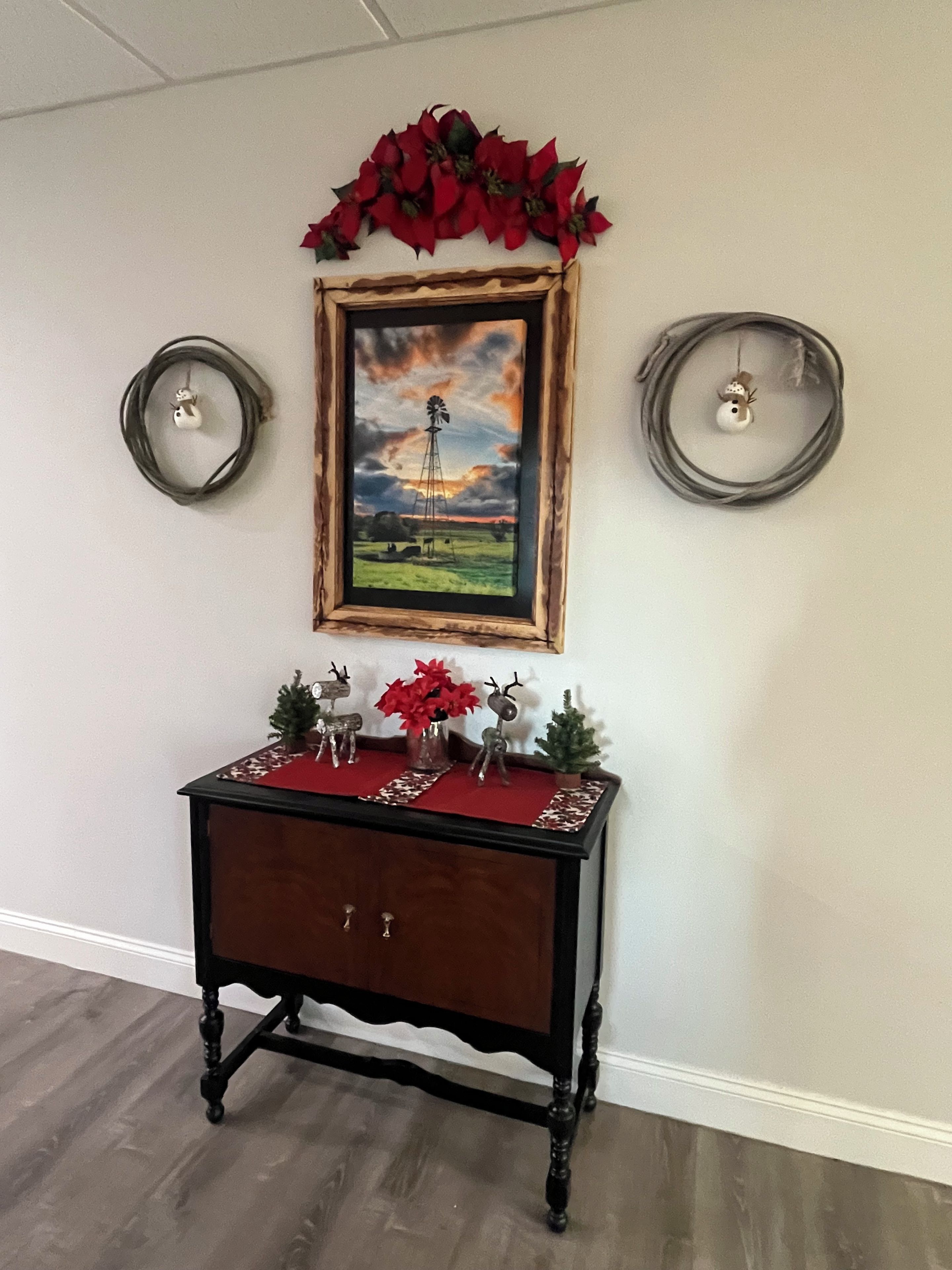 Una consola de madera con un mantel rojo, decorada con figuras de renos y un jarrón de flores rojas, ubicada frente a una pared con un cuadro enmarcado encima.